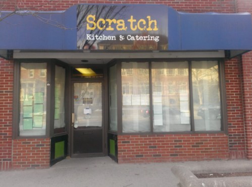 Exterior of Scratch Kitchen & Catering, featuring a blue awning and large windows with menus displayed.