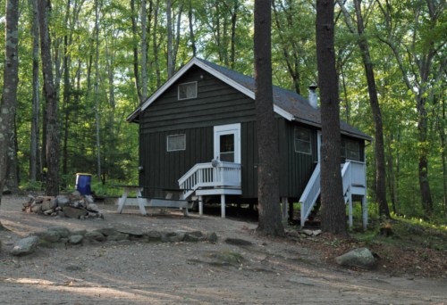 A small dark green cabin surrounded by trees, with a stone fire pit in front and a porch with steps.