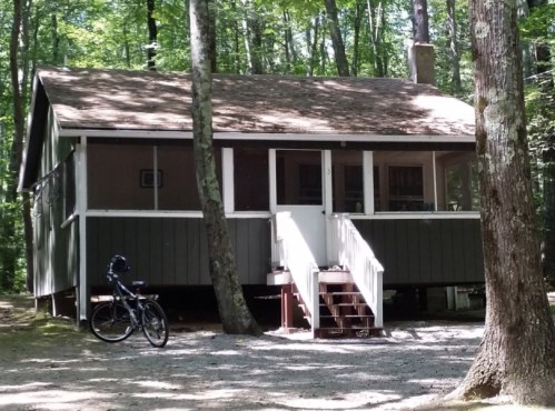 A cozy cabin in a wooded area, featuring a front porch and a bicycle parked nearby.