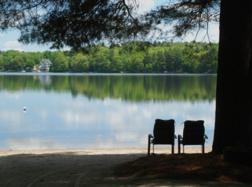 Two empty chairs by a serene lake, surrounded by trees and reflecting calm water under a clear blue sky.