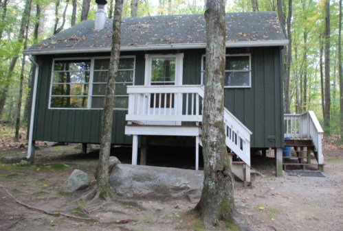 A small green cabin in the woods, surrounded by trees, with a white porch and steps leading to the entrance.
