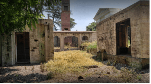 Abandoned buildings with crumbling walls and overgrown grass under a clear blue sky.