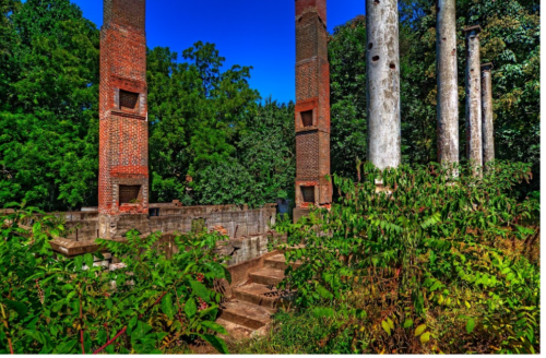 Ruins of brick pillars and stone foundations surrounded by lush greenery under a clear blue sky.