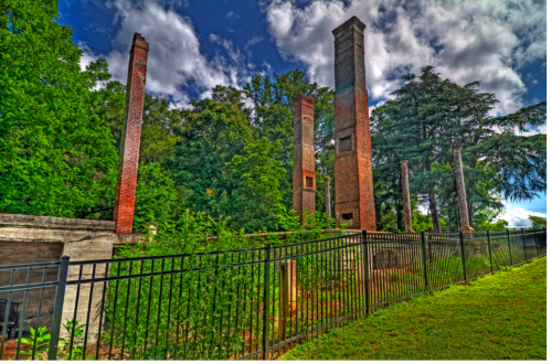Ruins of brick structures surrounded by greenery and a black fence under a cloudy sky.
