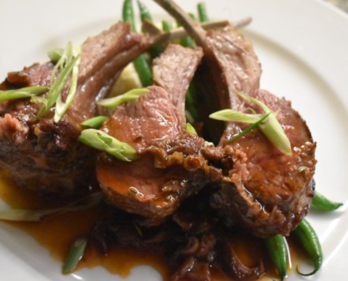 A plate of succulent lamb chops garnished with green onions, served on a bed of green beans and drizzled with sauce.
