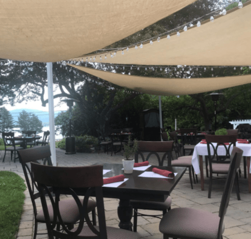 Outdoor dining area with tables set under a canopy, surrounded by greenery and a view of a lake in the background.