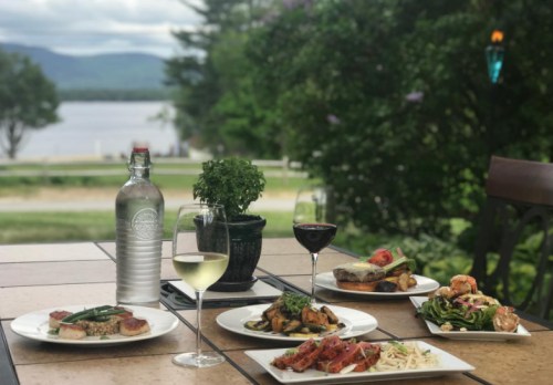 A scenic outdoor dining setup featuring various gourmet dishes, wine, and a view of a lake and mountains.