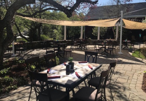Outdoor dining area with tables set for guests, surrounded by greenery and shaded by fabric canopies.