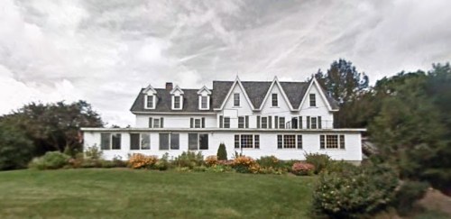 A large white house with multiple gables, surrounded by a lush green lawn and colorful flower beds under a cloudy sky.