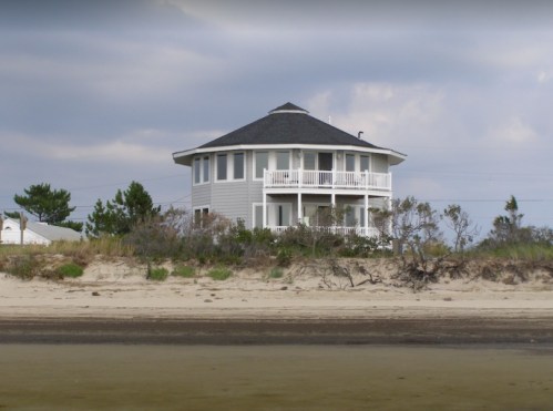 A round, two-story beach house with a gray roof, surrounded by sand and sparse vegetation.