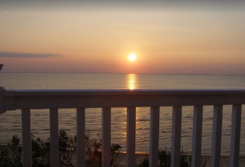 A serene sunset over calm waters, viewed from behind a white railing, with soft colors in the sky.
