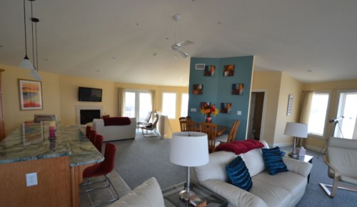 Bright, open living space with a kitchen, dining area, and cozy seating, featuring large windows and modern decor.