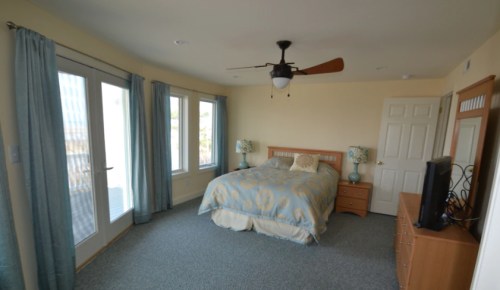 A spacious bedroom with a queen bed, light blue curtains, a ceiling fan, and a dresser with a TV.