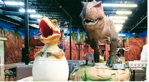 Two large dinosaur sculptures in a themed indoor space, one hatching from an egg and the other standing menacingly.