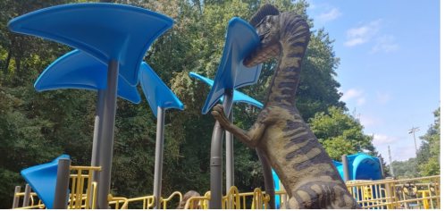 A dinosaur sculpture interacts with blue playground equipment under a sunny sky and trees in the background.