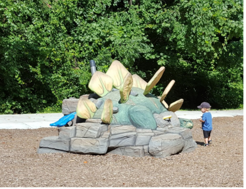 A child stands next to a large dinosaur sculpture in a playground, surrounded by trees and mulch.