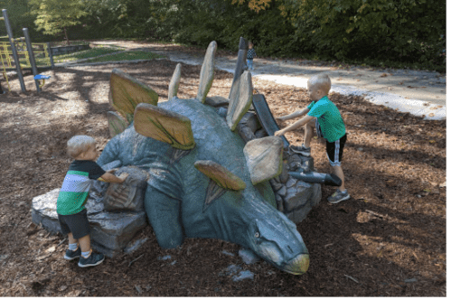 Two children play on a dinosaur sculpture at a park, interacting with its textured surface in a natural setting.