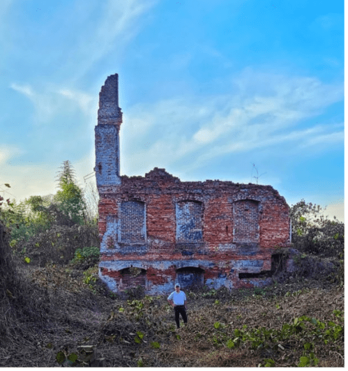 A person stands in front of a partially collapsed brick building surrounded by overgrown vegetation under a blue sky.