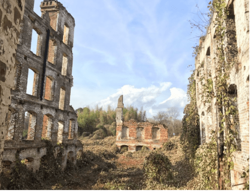 Ruins of a brick building overgrown with vegetation, under a blue sky with scattered clouds.