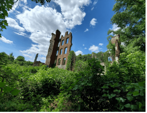Ruins of a brick building surrounded by lush greenery and under a bright blue sky with fluffy clouds.