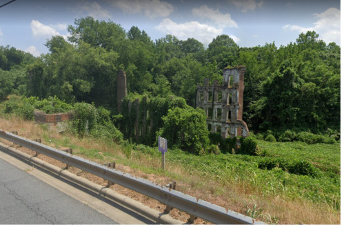 Overgrown ruins of a brick structure surrounded by dense greenery, located near a roadside.