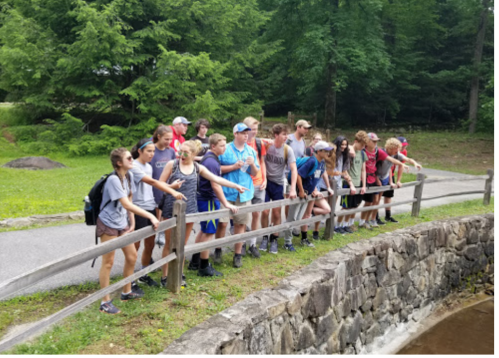 A group of young people stands by a wooden fence, looking at a body of water in a forested area.