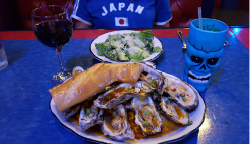 A plate of oysters topped with sauce, a side salad, a blue skull cup, and a glass of red wine on a blue table.