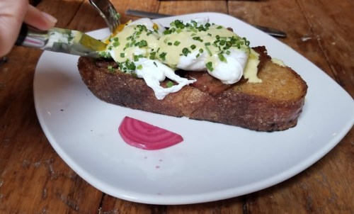 A slice of toasted bread topped with poached eggs, hollandaise sauce, and chives, served on a white plate with pickled onions.