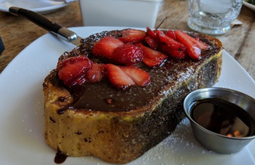A slice of French toast topped with fresh strawberries and chocolate sauce, served with a small cup of syrup.
