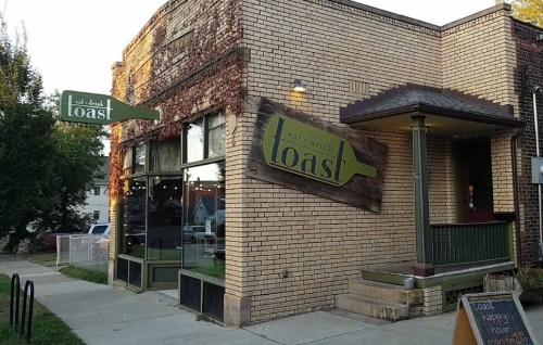 Exterior of a brick building with ivy, featuring a sign that reads "eat drink toast" and a sidewalk menu board.