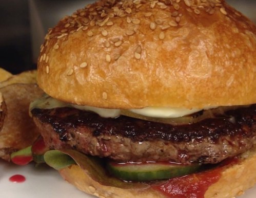 Close-up of a sesame seed bun burger with a juicy patty, pickles, and sauces, served with crispy potato chips.