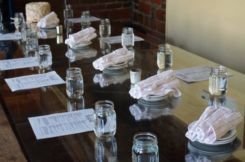 A dining table set with plates, folded napkins, water jars, and menus, against a brick wall backdrop.