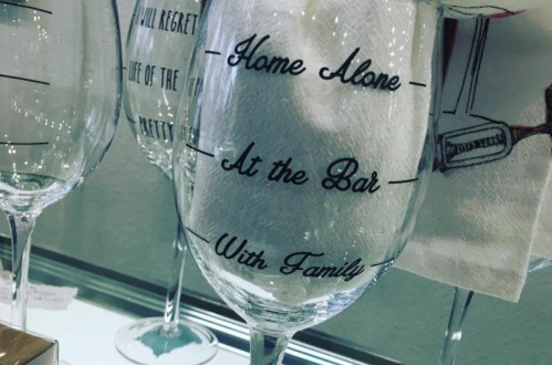A close-up of a wine glass with playful text: "Home Alone, At the Bar, With Family."