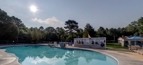 A sunny day at a swimming pool surrounded by trees, with a small building and lounge chairs nearby.