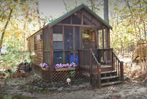 A cozy wooden cabin with a green roof, surrounded by trees and flowers, featuring a porch and steps leading up to the entrance.