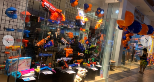 A display of colorful toy blasters and accessories in a store, with balloons and decorations in the background.
