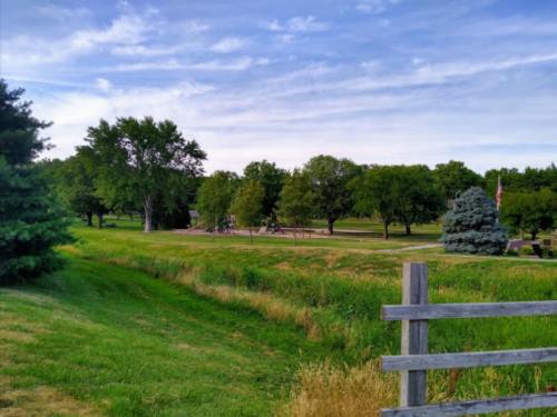 A serene landscape featuring green fields, trees, and a wooden fence under a blue sky with scattered clouds.