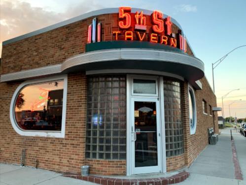 Exterior of 5th St Tavern, featuring a neon sign, brick facade, and large glass windows at sunset.