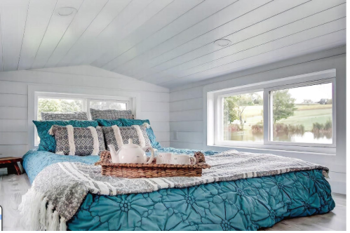 Cozy bedroom with a large bed, decorative pillows, and a view of a serene landscape through two windows.