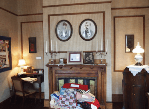 A vintage room featuring a fireplace, framed portraits, a desk, and a stack of colorful quilts.