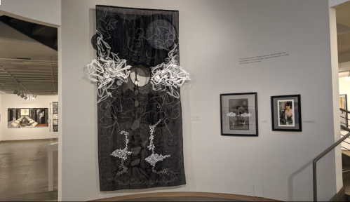 A large textile artwork hangs on a gallery wall, flanked by two framed images, in a modern art exhibition space.