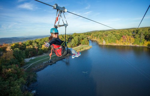 A person zip-lining over a scenic lake surrounded by colorful autumn trees under a clear blue sky.