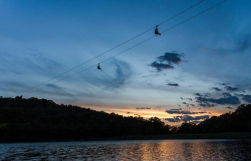 Two zipliners glide over a lake at sunset, with a colorful sky and silhouettes of trees in the background.