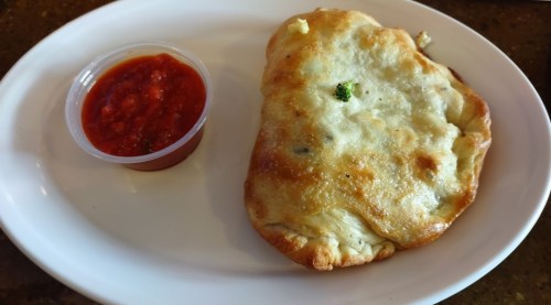 A golden-brown stuffed pastry on a plate, served with a small cup of red dipping sauce.