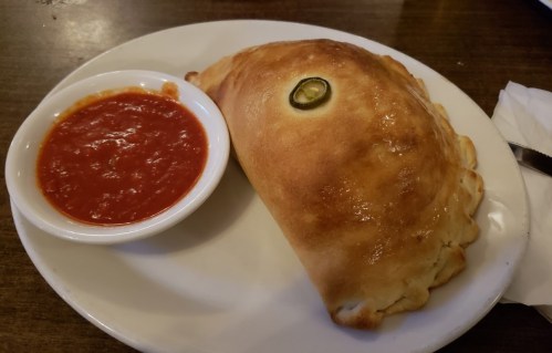 A golden-brown calzone on a plate, served with a side of marinara sauce and a slice of jalapeño on top.