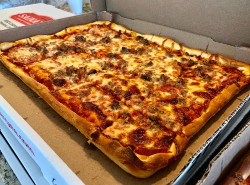 A rectangular pizza with cheese and sausage, served in a cardboard box.