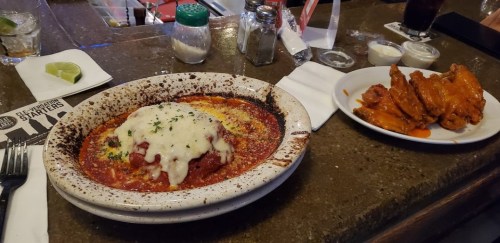 A plate of cheesy baked pasta with marinara sauce next to a serving of crispy chicken wings on a restaurant table.