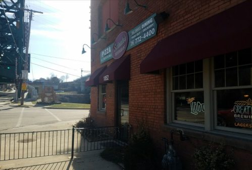 Exterior of a pizza restaurant with a red brick facade, awnings, and a sign displaying the name and contact number.