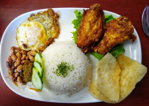 A plate of rice, fried chicken wings, a fried egg, beans, cucumber slices, and crispy crackers on a bed of lettuce.
