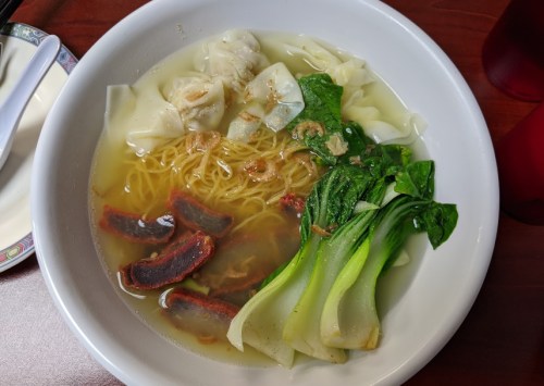 A bowl of soup with wontons, noodles, bok choy, and slices of meat in a clear broth.